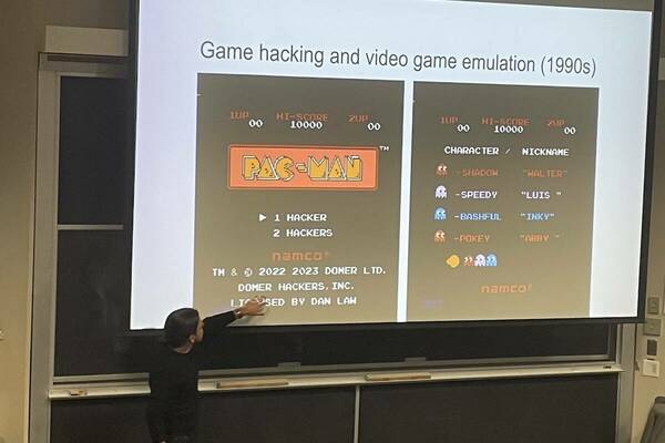 Luis Felipe Murillo points at a large classroom screen showing images of the title screens from the Pac-Man video game with certain words altered via hacks